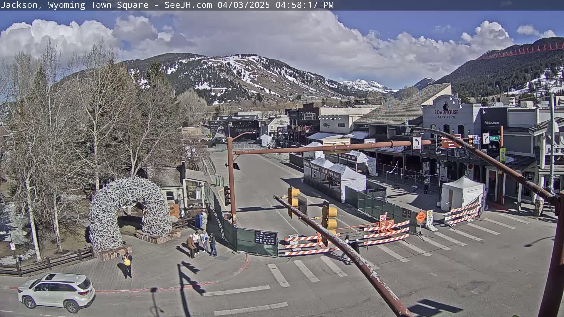 A town square with snow-capped mountains in the background under partly cloudy skies, featuring several buildings, a temporary road closure, and a large antler arch.
