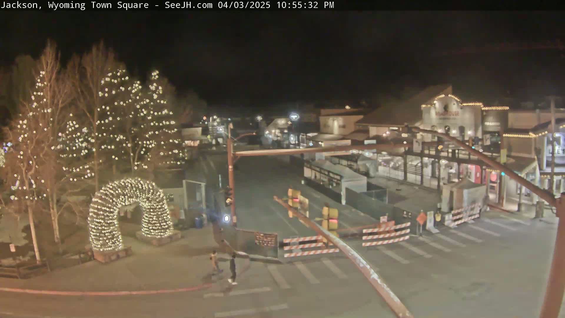 A nighttime view of a town square with a light-covered archway, buildings, and construction barriers.