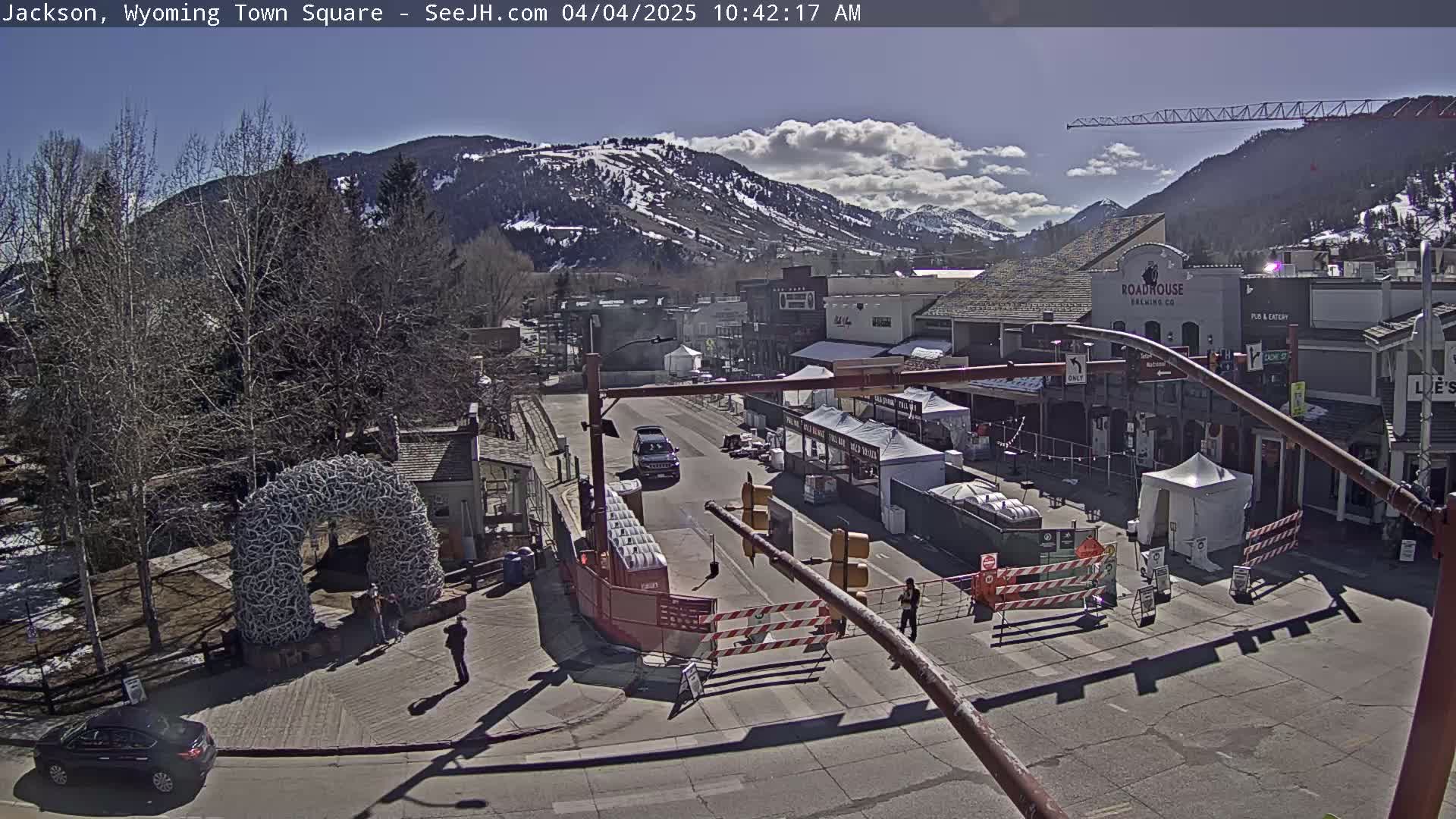 A partly sunny day shows a town square with several buildings, a large antler arch, and temporary stalls or tents set up in the street, all set against a backdrop of snow-capped mountains.