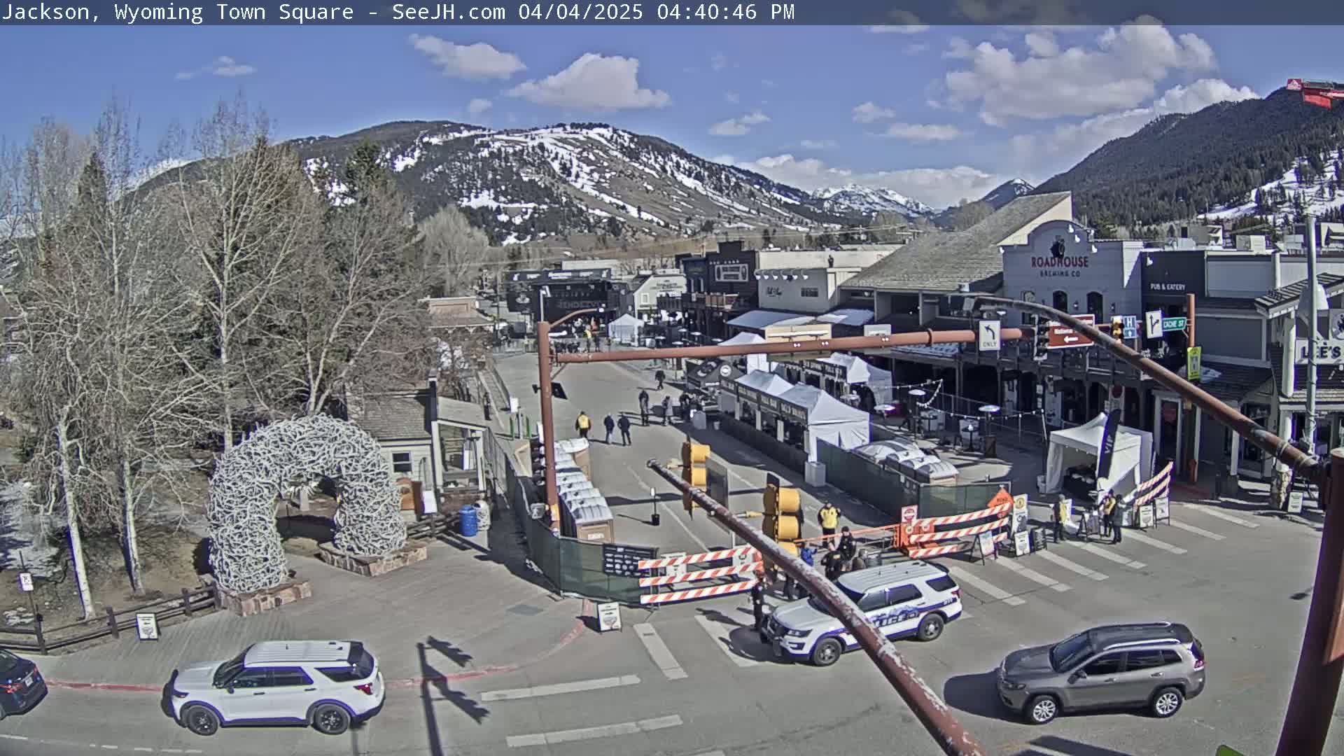 A town square with a large antler arch, numerous temporary vendor stalls, and several vehicles, all under a mostly sunny sky with some clouds and snow-capped mountains in the background.
