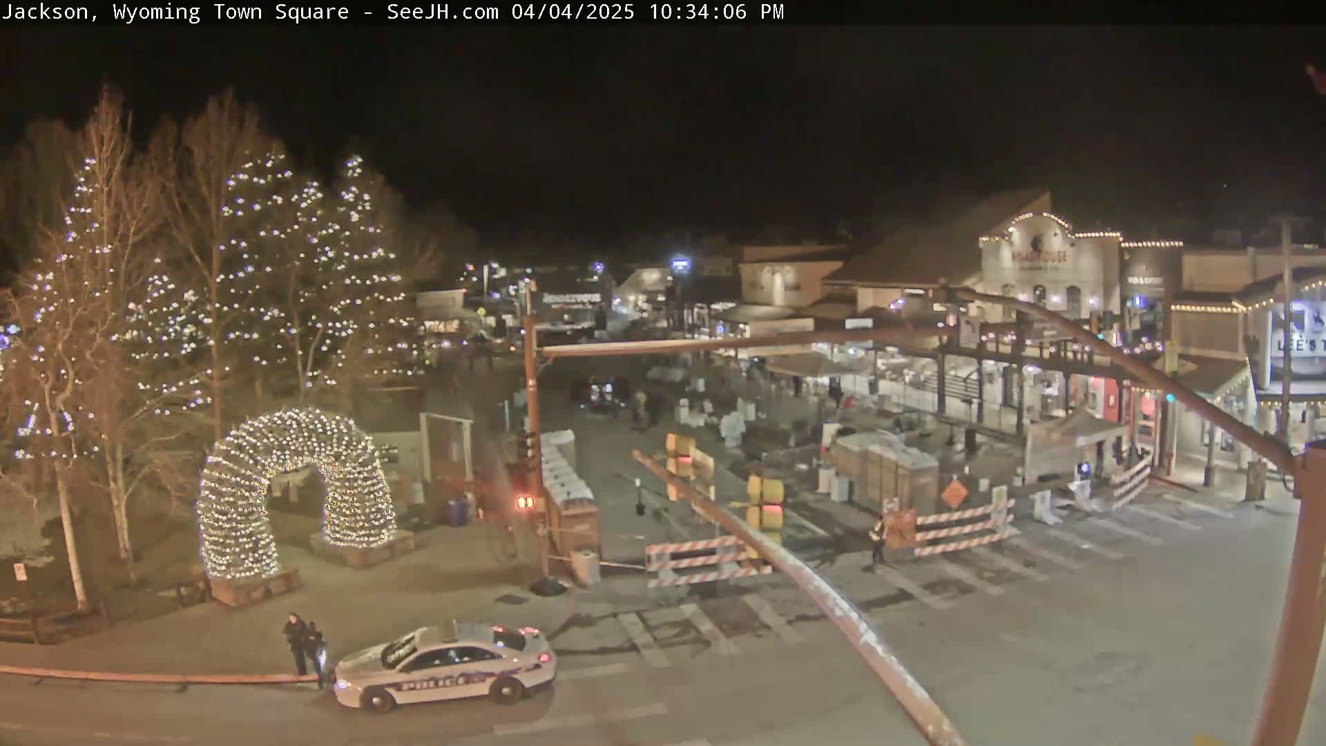 A nighttime view of a town square with Christmas lights illuminating trees and an archway, a police car parked near two officers, and construction barriers blocking off parts of the street.