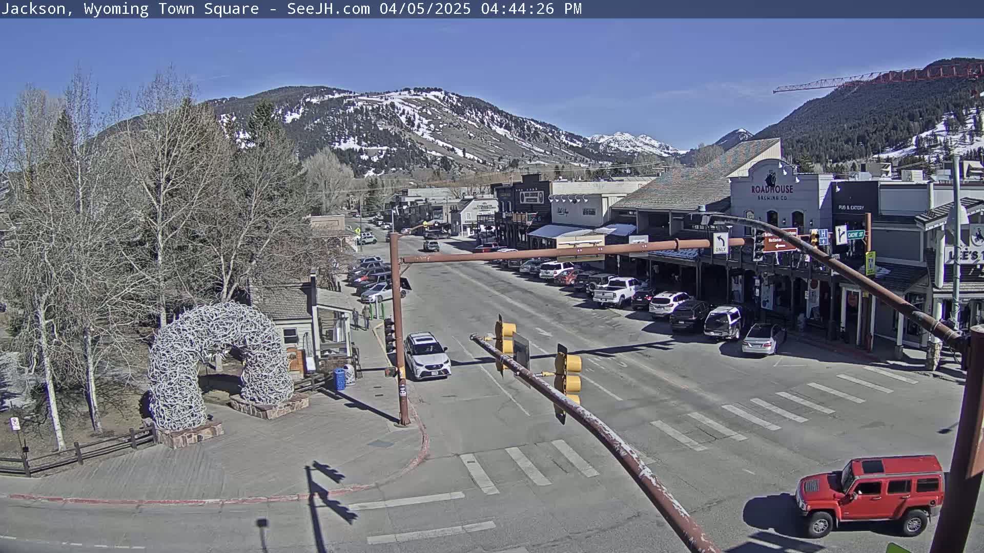 A sunny view of a town square, featuring a street with parked cars and buildings, a large antler arch, and snow-capped mountains in the background.