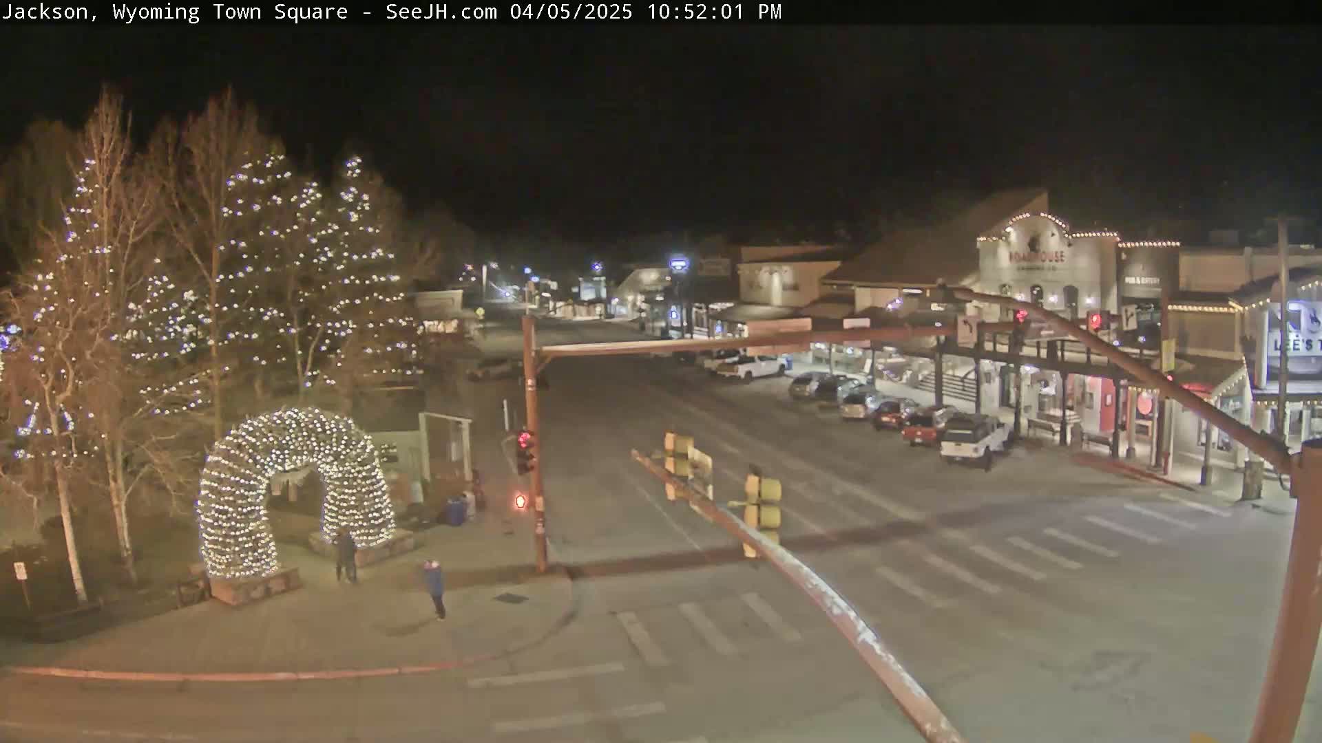 A nighttime, snow-covered town square is illuminated by streetlights and Christmas lights strung on trees and an archway; several cars are parked along the sides of the road.