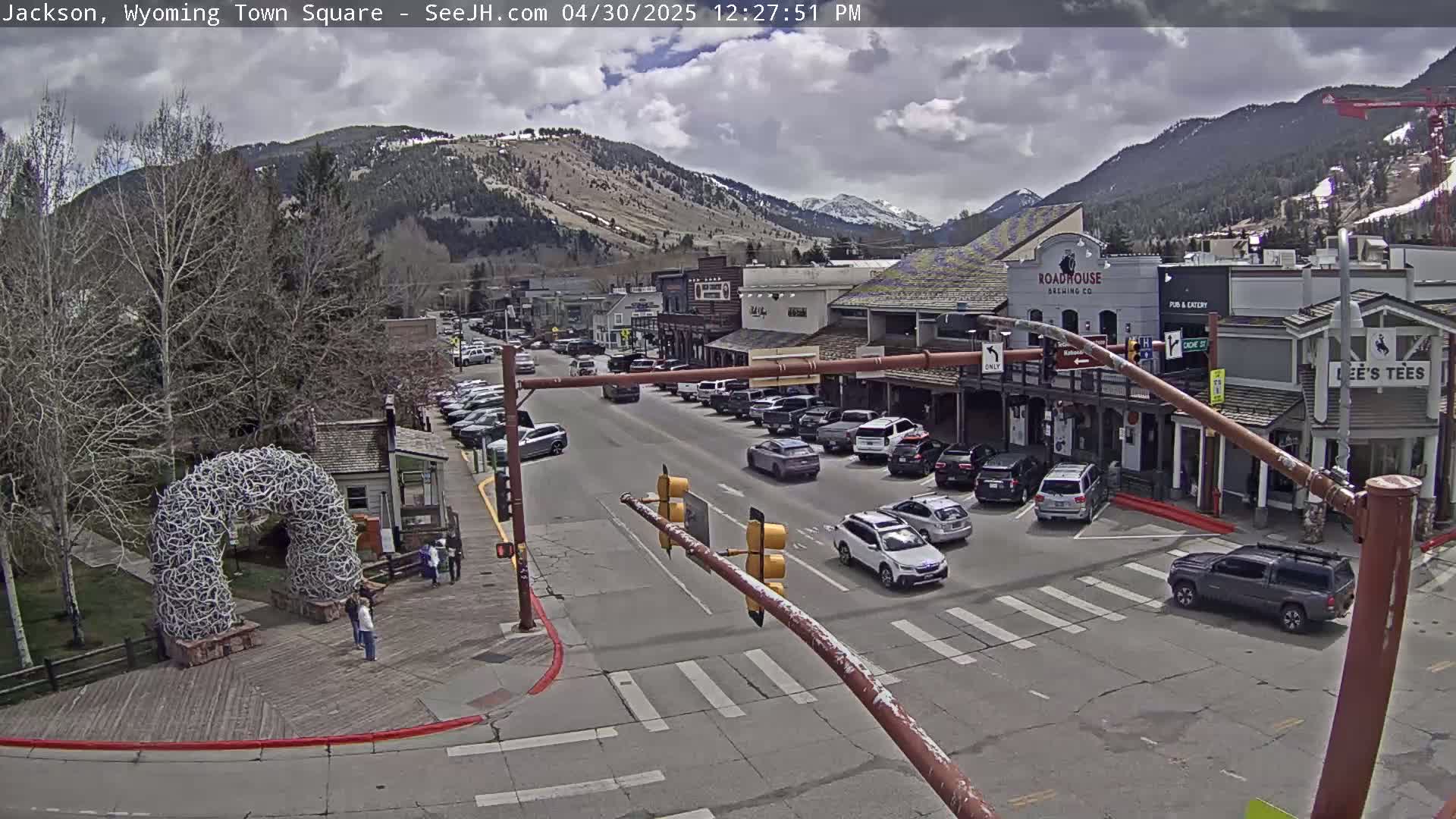A town square with parked cars and buildings, under a partly cloudy sky, with mountains in the background.