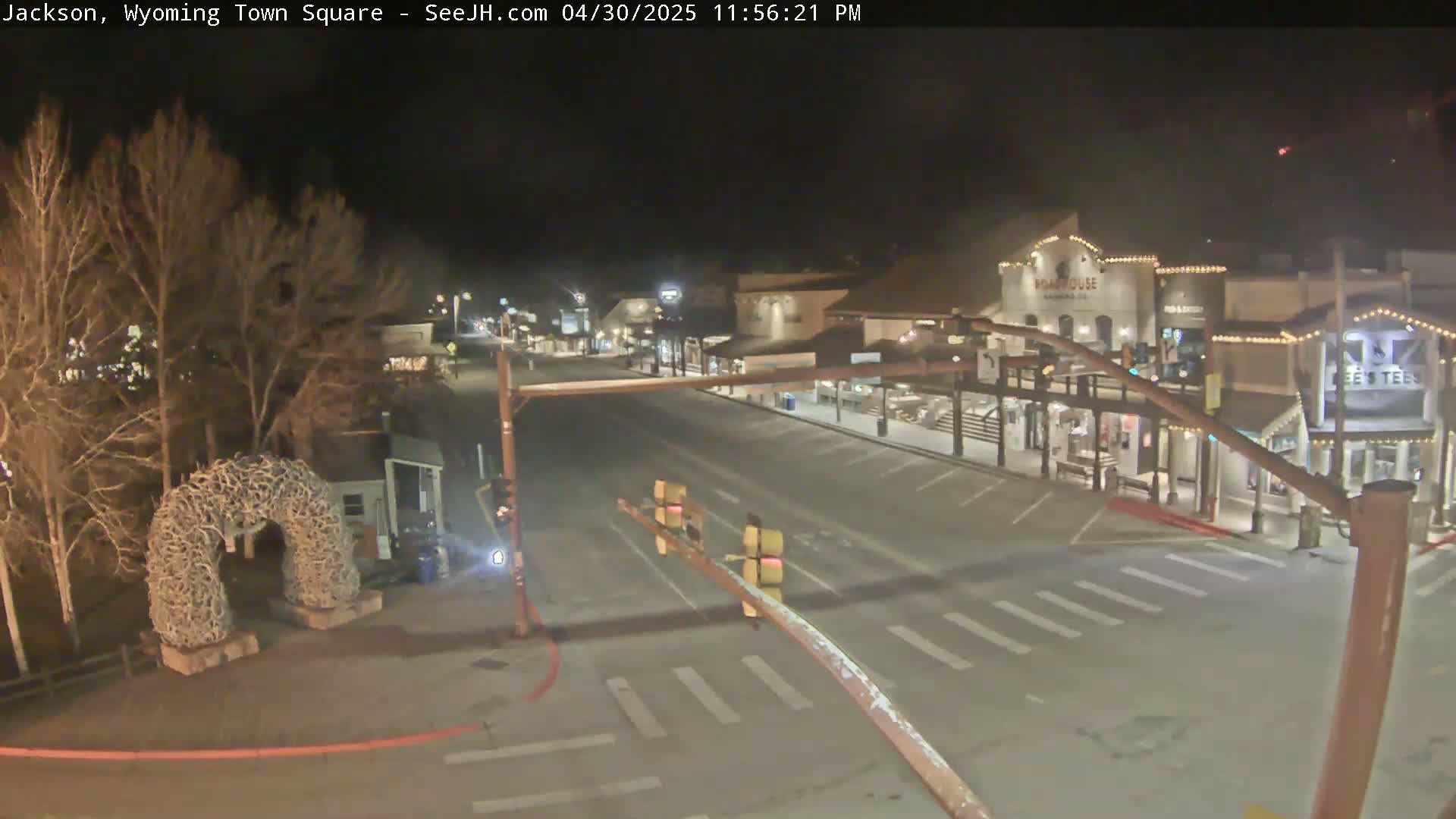 A nighttime view of a mostly empty town square with bare trees, a large antler arch, and several buildings with lights on.