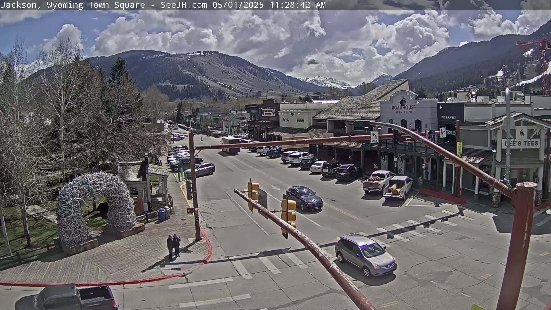 A partly cloudy day in a mountain town square, showing parked cars along a street lined with buildings, and a large antler arch in a nearby park.