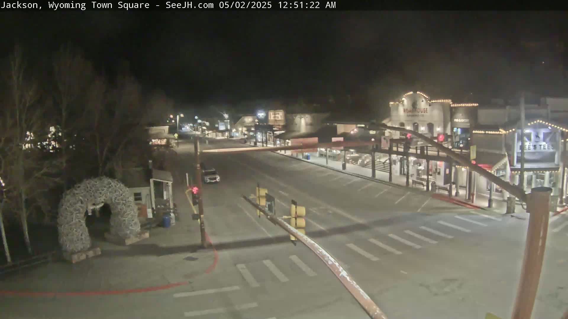 A nighttime view of a mostly empty town square, featuring a vehicle, buildings with lights, and a large antler arch.