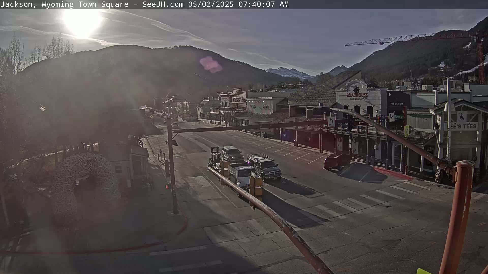 A sunny morning view of a town square with several vehicles on a street in front of buildings nestled against a mountain backdrop.