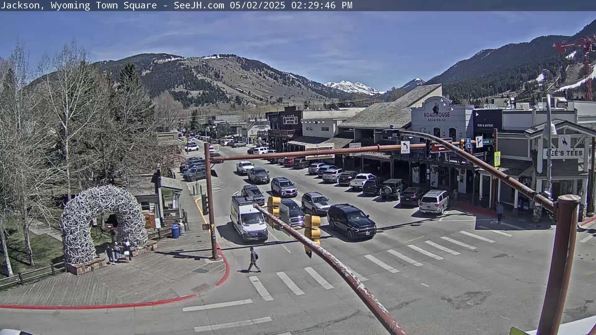 Under a clear blue sky, a town square shows several vehicles at an intersection in front of buildings, with a large antler arch visible to the lower left.