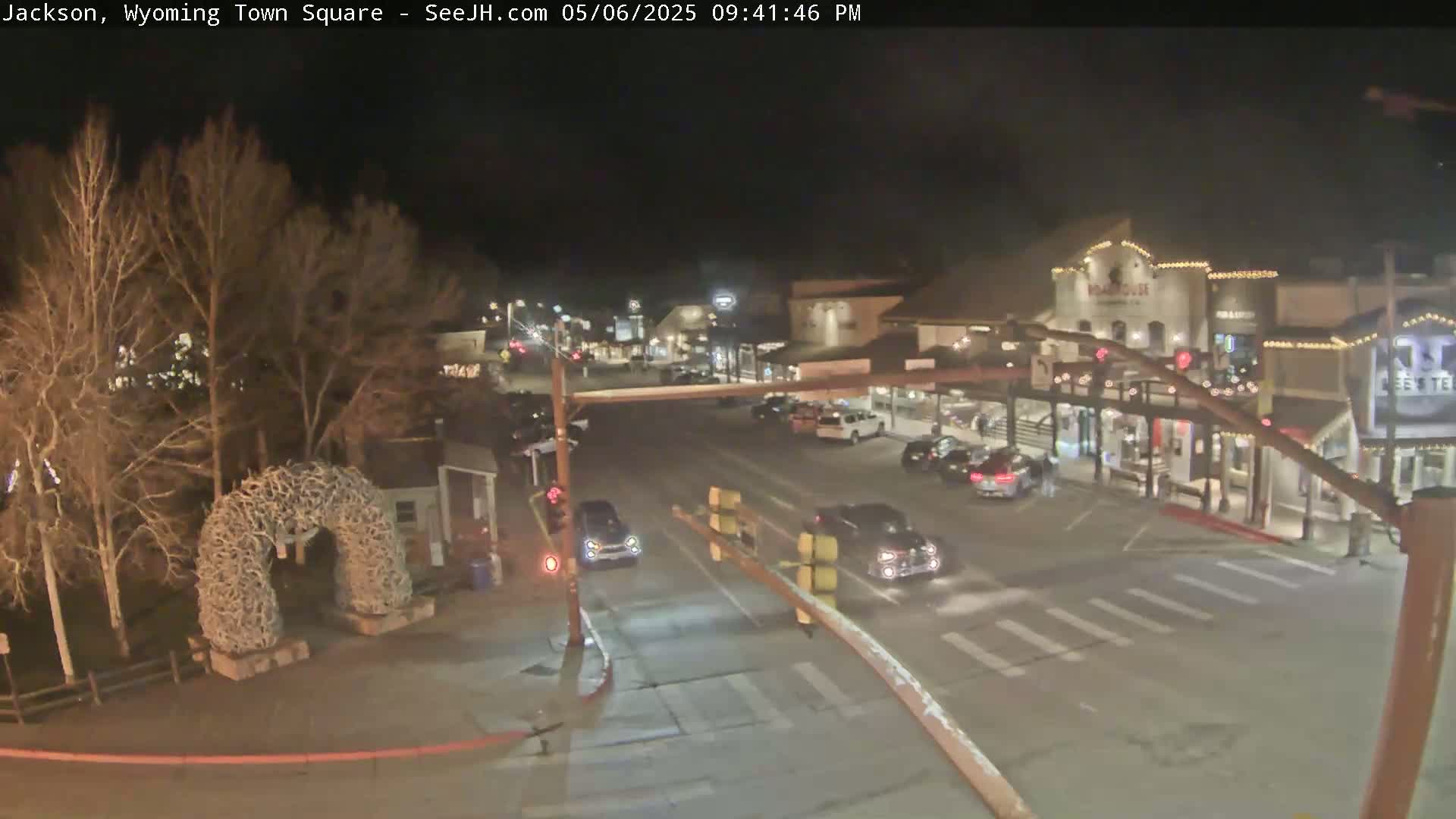 A nighttime view of a town square with several cars at an intersection, buildings with lights, and bare trees, under a clear dark sky.