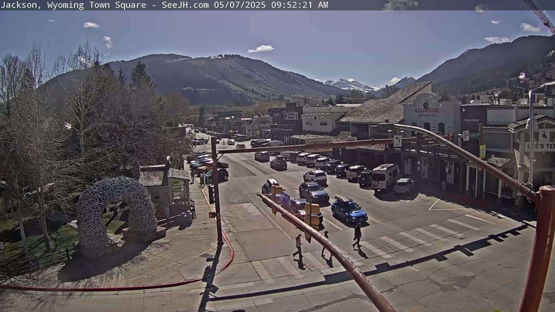 A sunny town square with cars parked along the street, pedestrians crossing, and mountains in the background.
