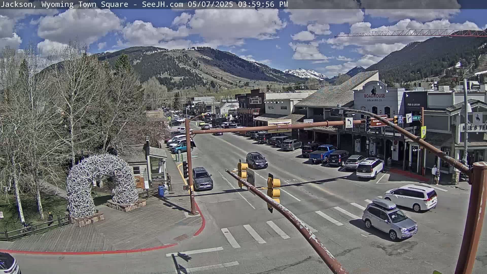 A sunny town square scene shows a street with cars, buildings, and a large antler archway, all nestled against a backdrop of mountains.