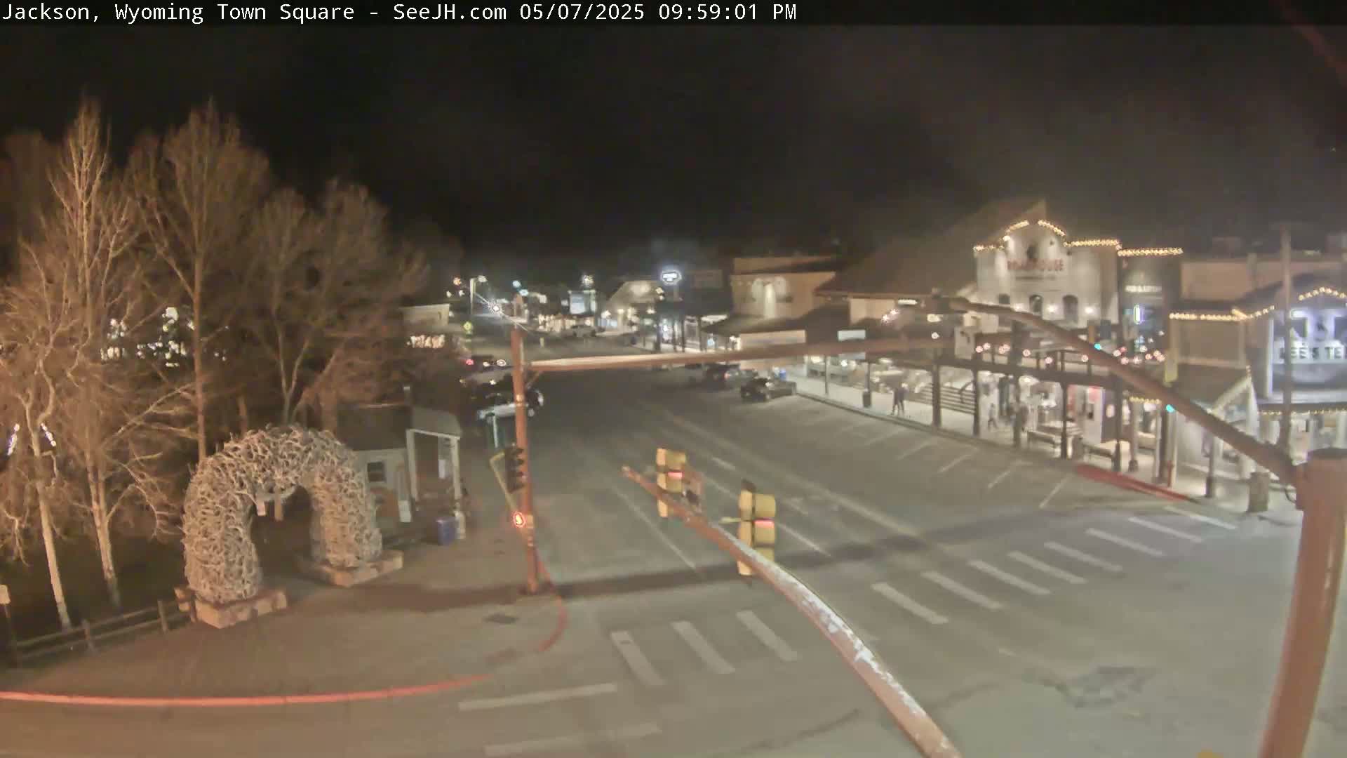 A nighttime view of a town square with a street intersection, buildings with lights, and bare trees.