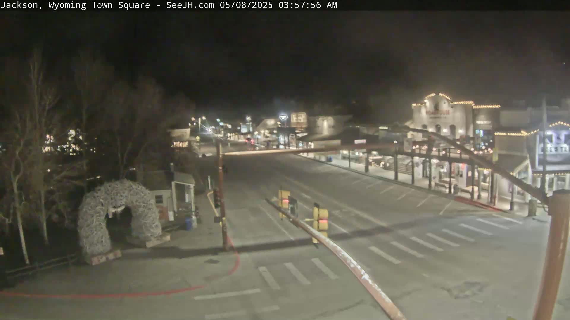 A mostly empty town square at night, with a few streetlights illuminating the roadway and buildings, and an antler archway visible in the foreground.