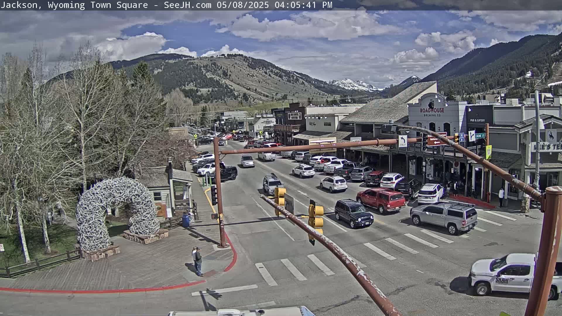 A partly cloudy afternoon scene shows a town square with cars driving on a street lined with buildings, a large antler arch, and mountains in the background.