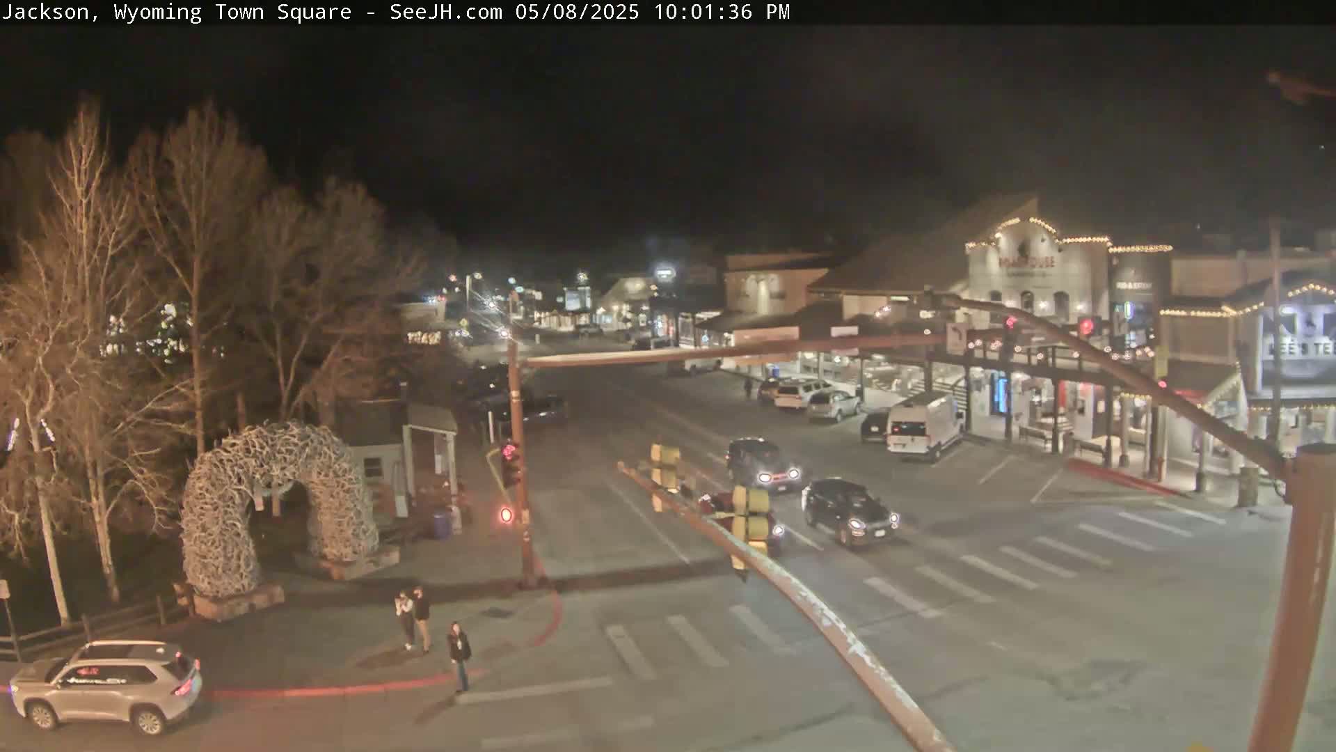 A nighttime view of a town square with several cars at an intersection, under a clear sky, featuring a large antler archway in the foreground.