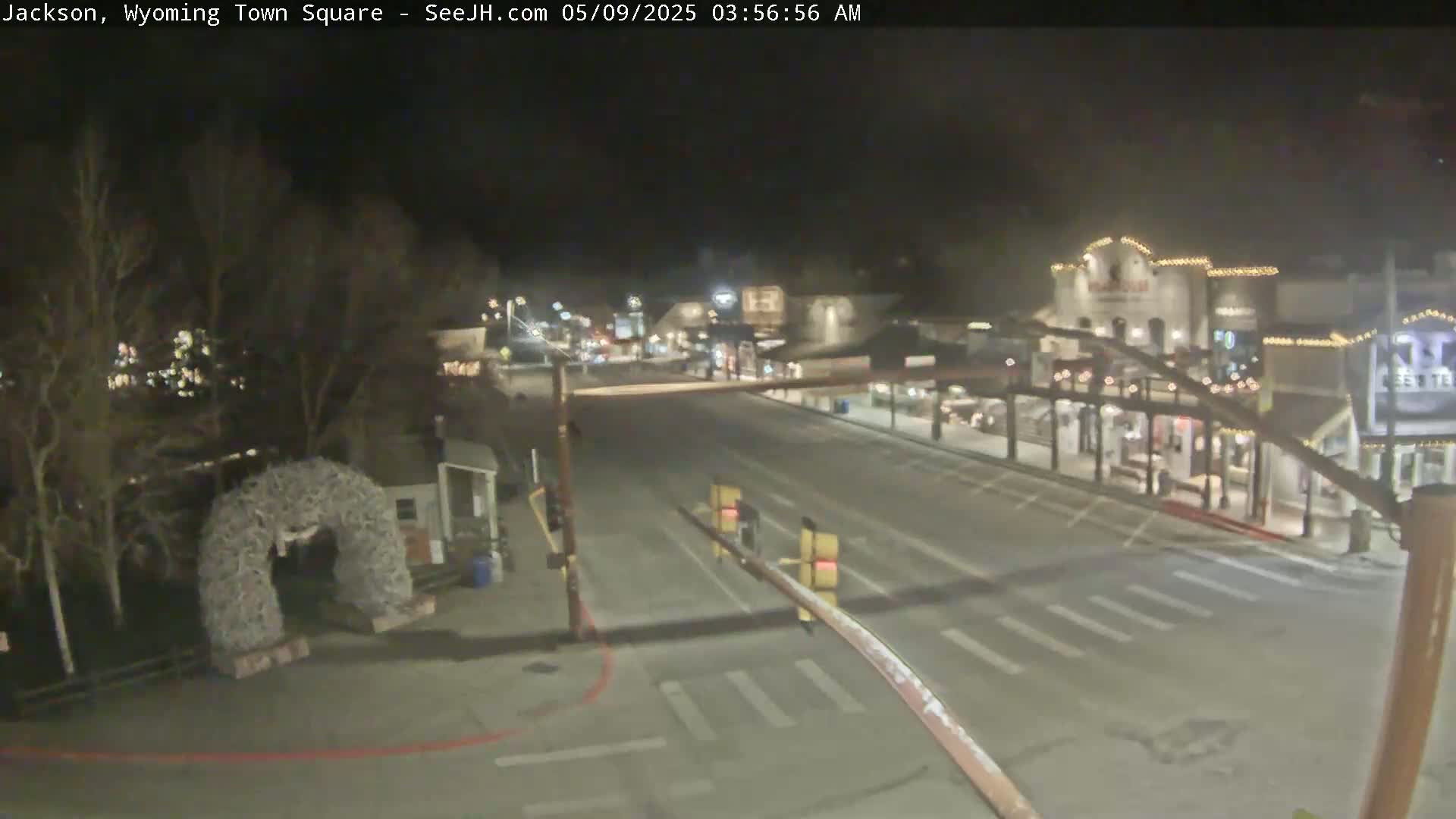 A nighttime, mostly empty town square with a large antler archway visible on the left and several buildings illuminated along the right side of a road.