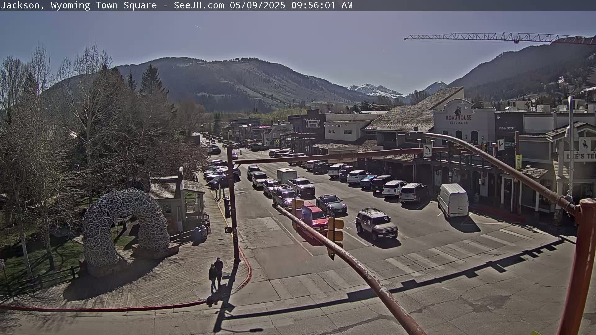 A sunny day reveals a town square with cars on the street, buildings lining the sides, and a large antler arch in a park-like setting, backed by mountains.