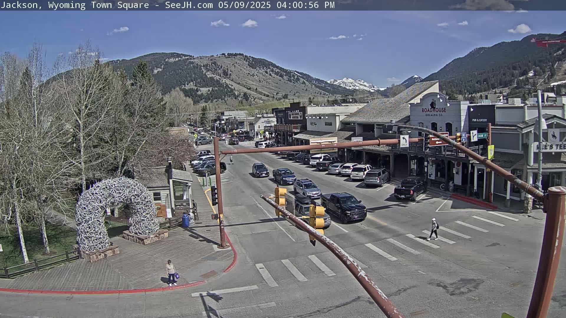 A sunny day in a town square shows cars parked and driving on a street lined with buildings, with a large antler arch in a nearby park.