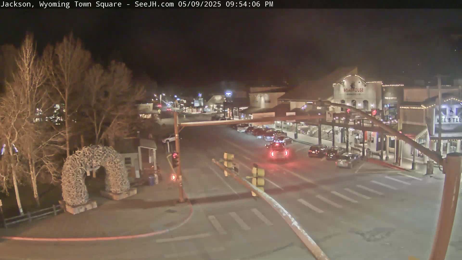 A nighttime, dimly lit town square features sparse traffic on a street that intersects with another road, passing in front of buildings with lights; a large antler arch is visible in the foreground.