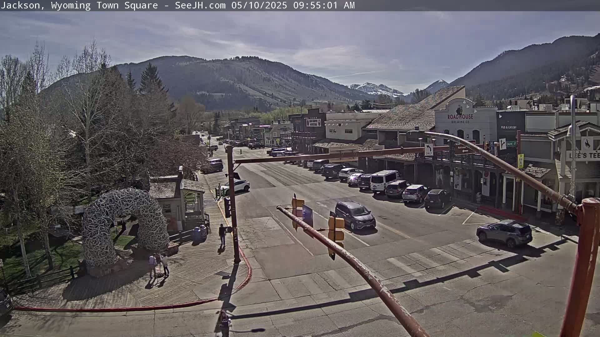 A sunny view of a town square, with cars parked along the street and a few pedestrians, nestled in a valley between mountains.