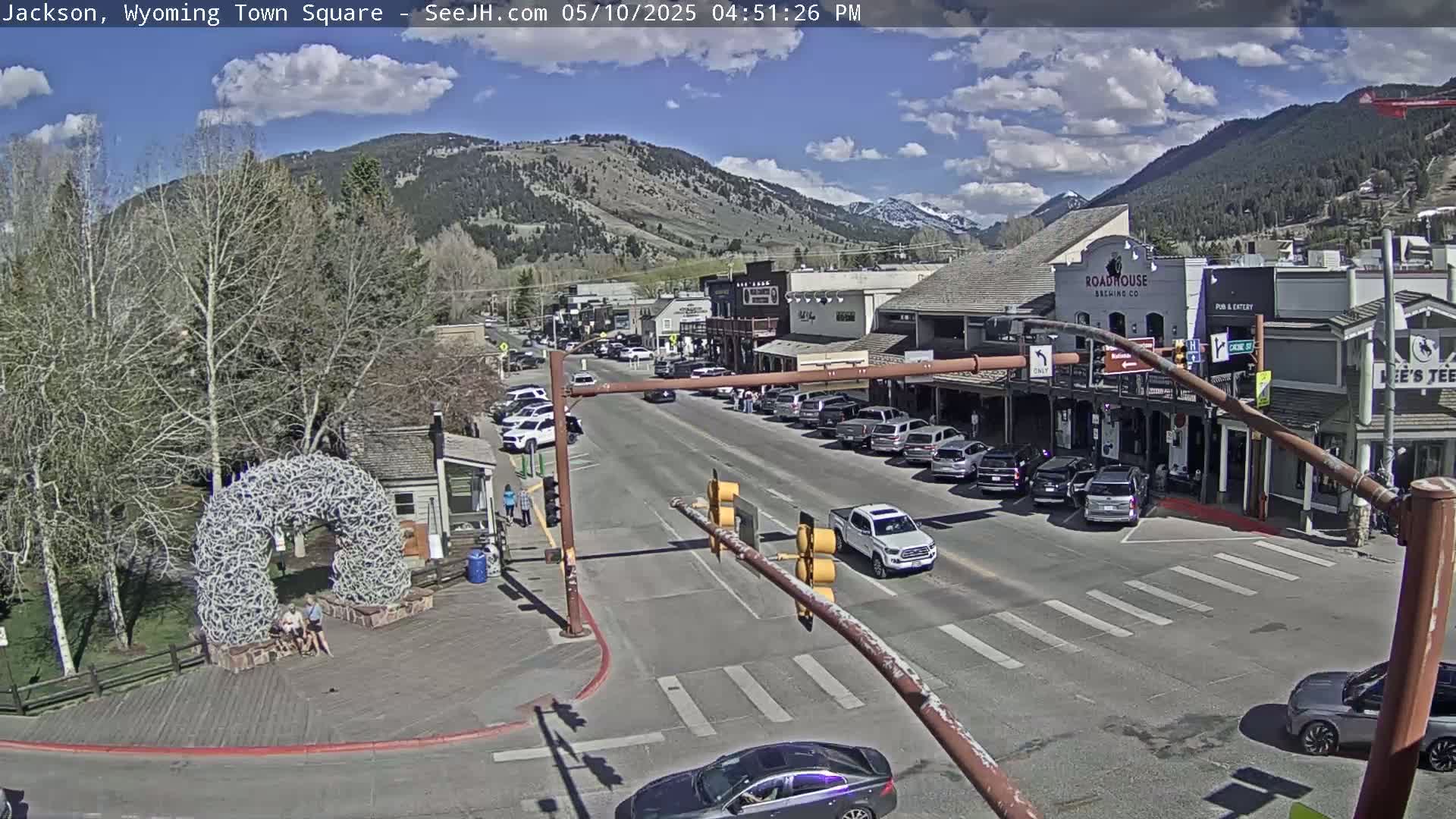 A town square with parked cars and buildings under a mostly sunny sky, with mountains in the background.