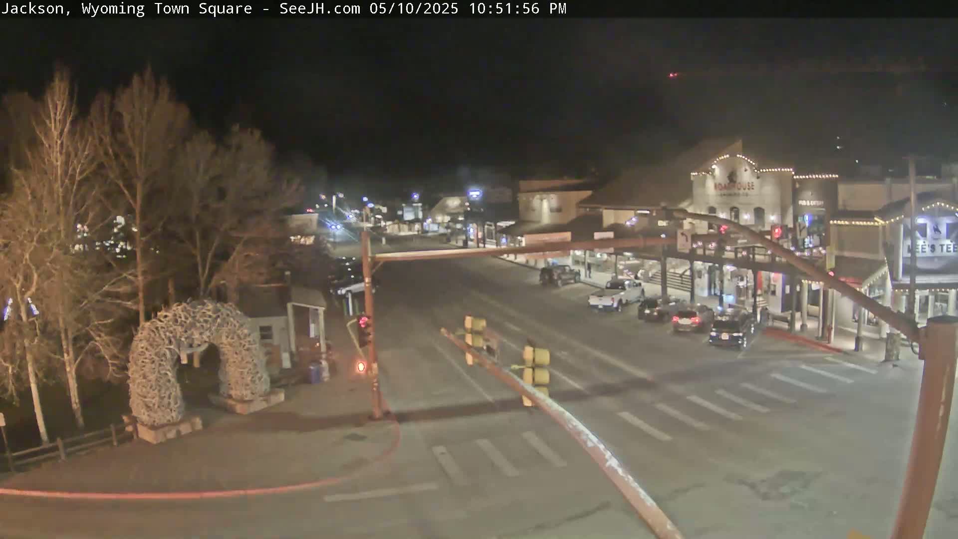 A nighttime, illuminated view of a town square with several vehicles parked and driving on the streets, passing by buildings and a large antler arch.
