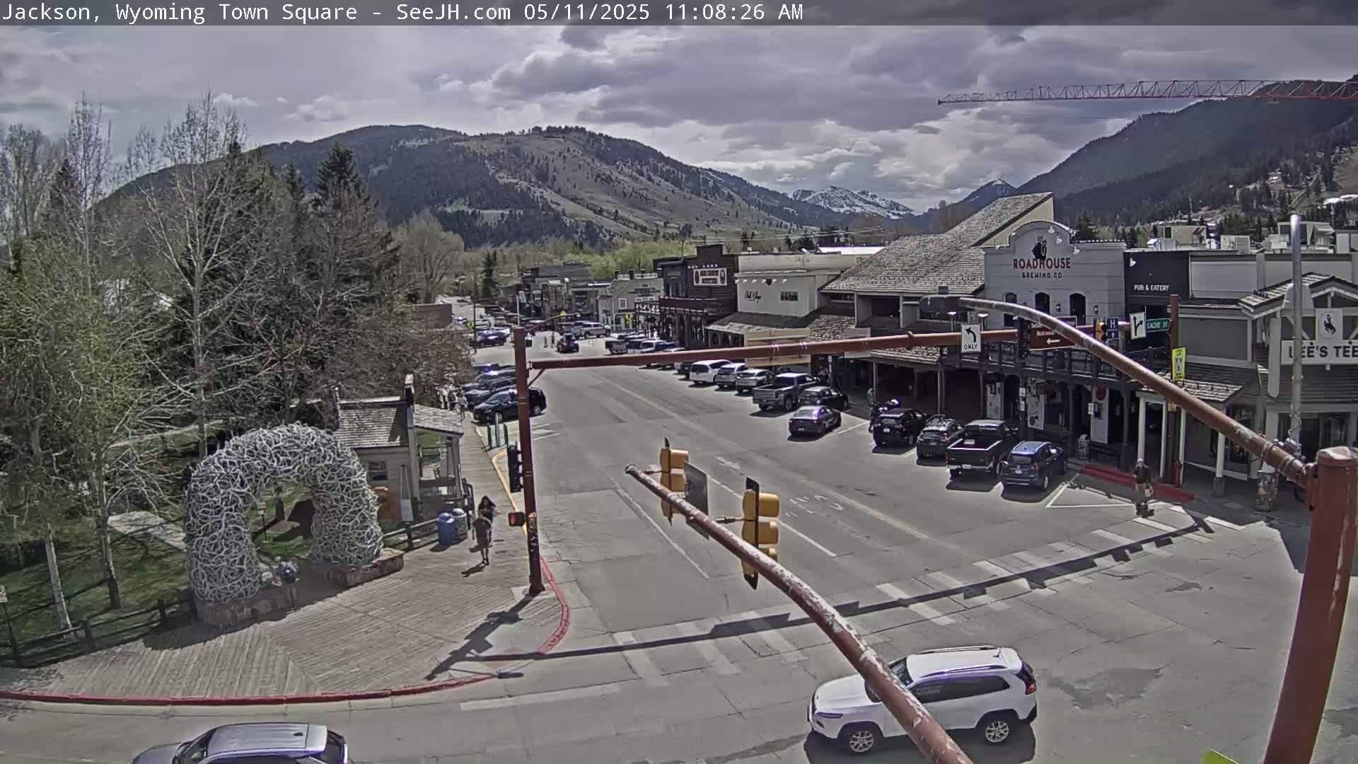 A partly cloudy day reveals a town square with parked cars, buildings, and pedestrians, situated at the base of a mountain range.