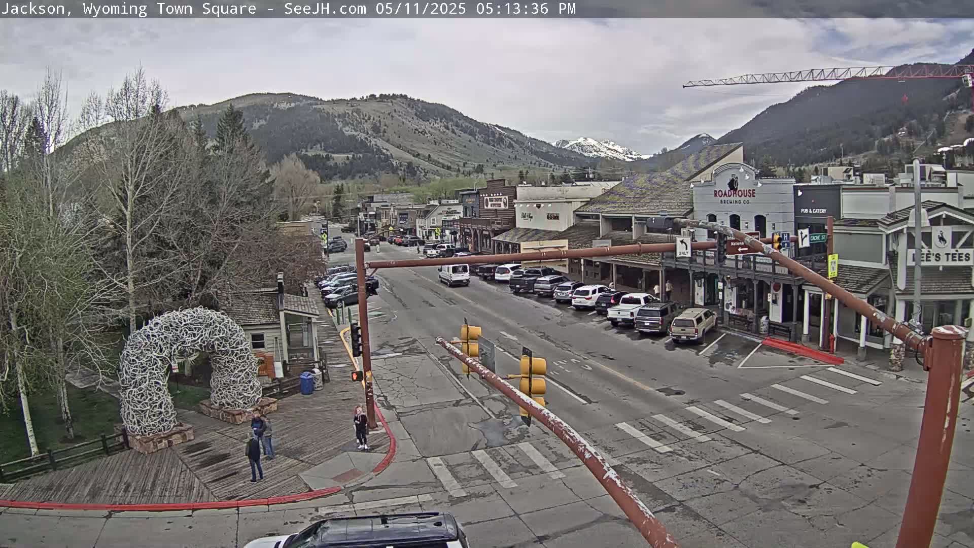 A town square with parked cars and a few pedestrians, under an overcast sky, is nestled at the foot of a mountain range.