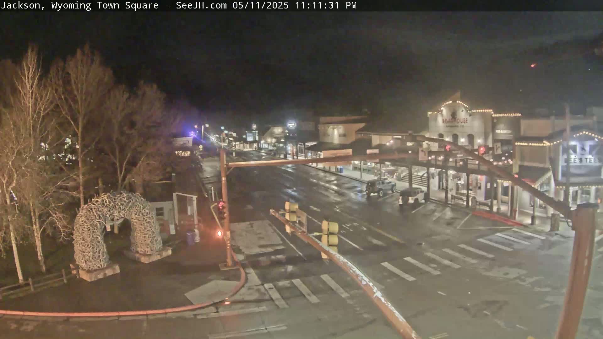 A wet nighttime scene shows a town square with sparse traffic, a large antler arch, and buildings with lights.