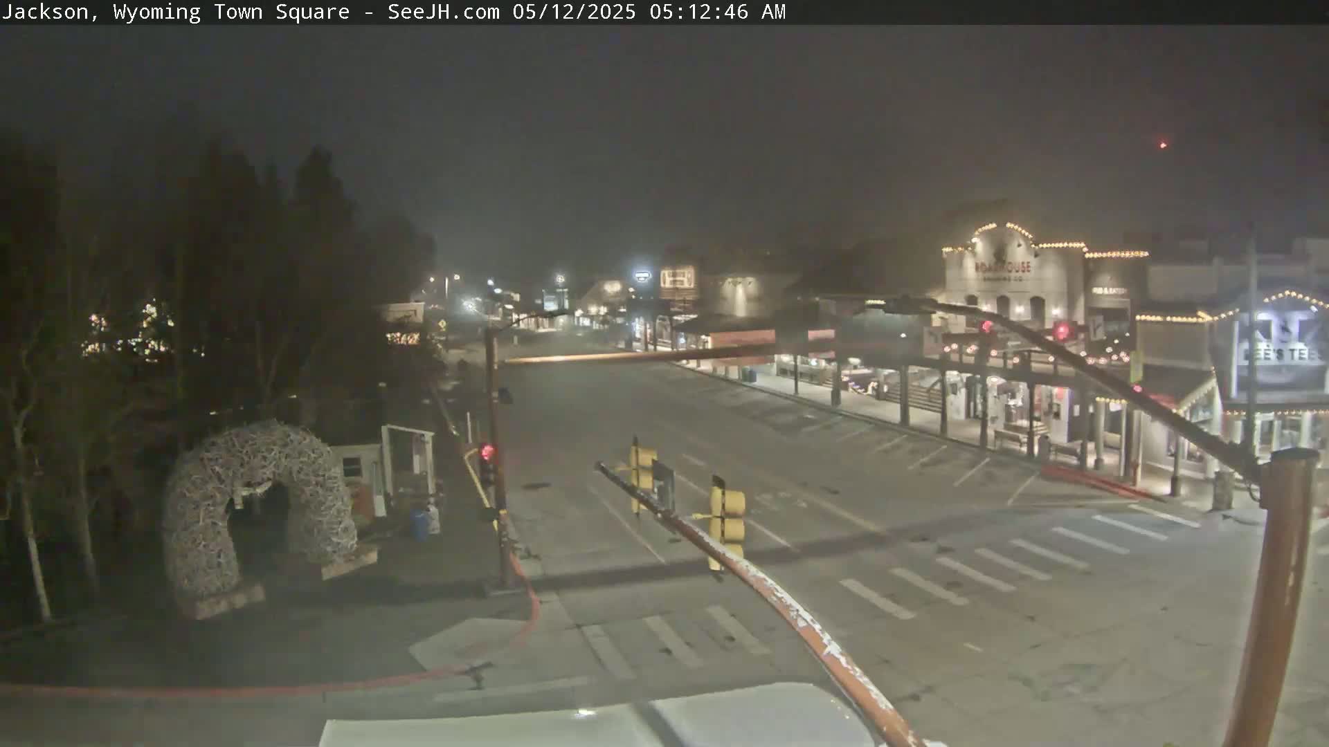 A mostly empty town square at night, shrouded in fog, shows several buildings with lights on and a decorative archway in the foreground.