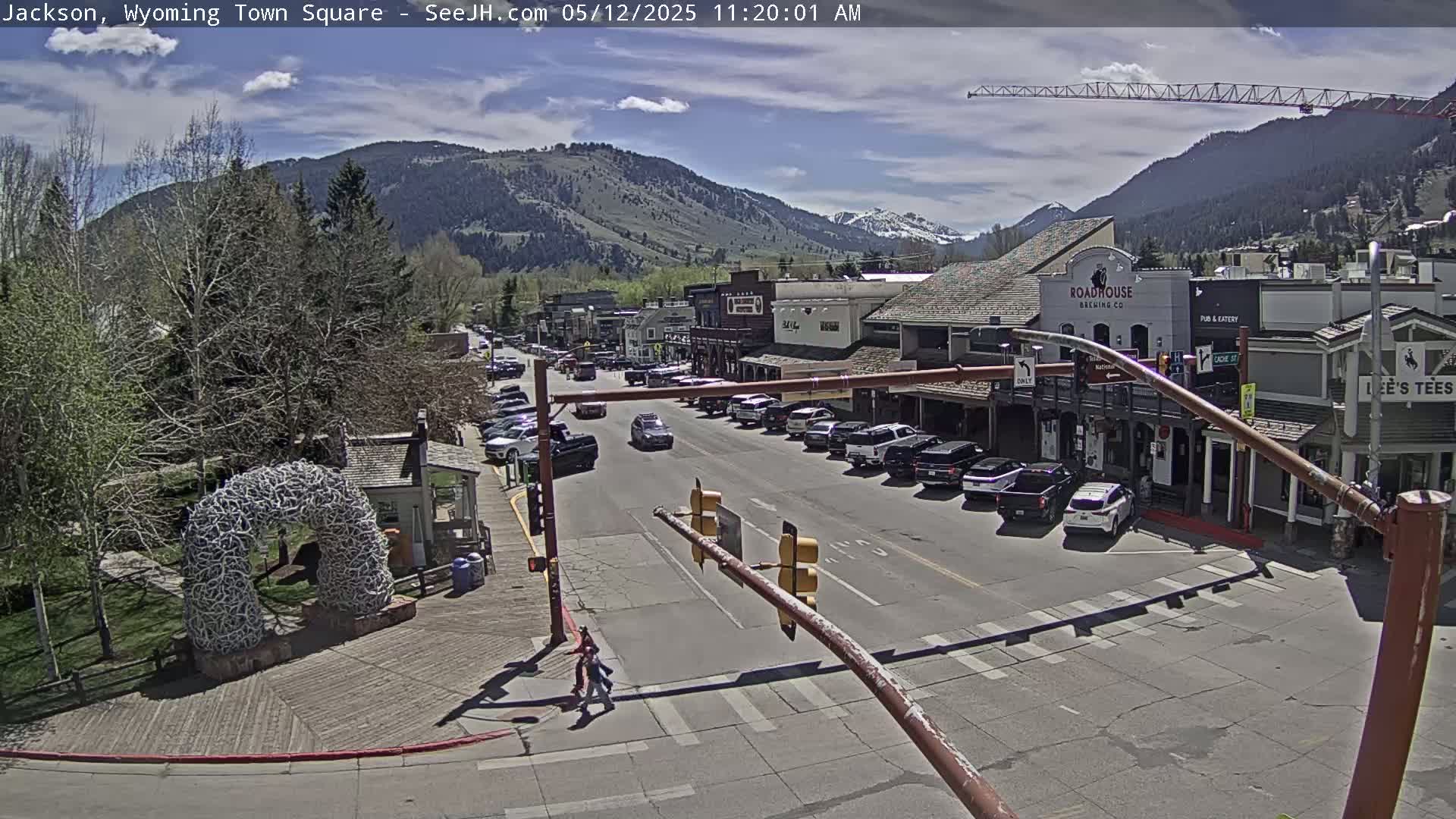 A sunny town square scene shows a street with parked cars and a few pedestrians,  bordered by buildings nestled at the base of a mountain range.