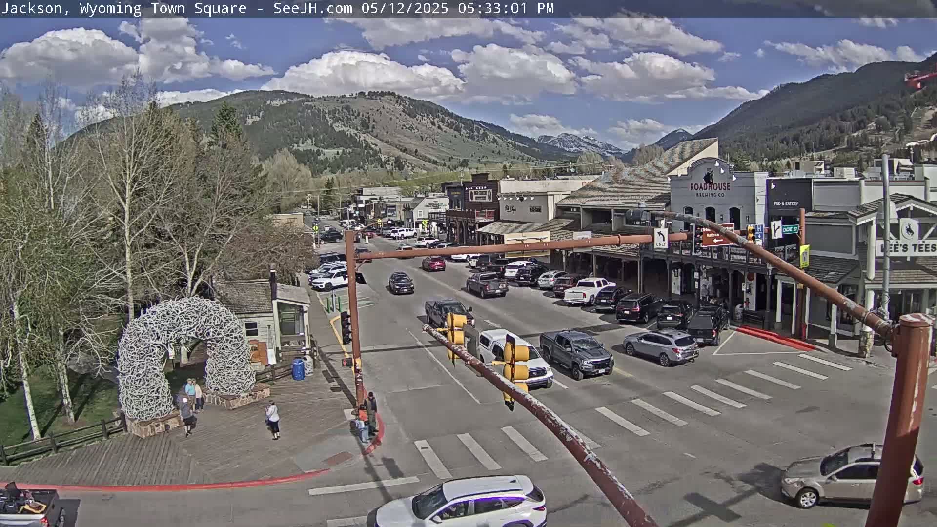 A partly cloudy day in a town square, showing a street with cars, buildings, and a large antler arch.