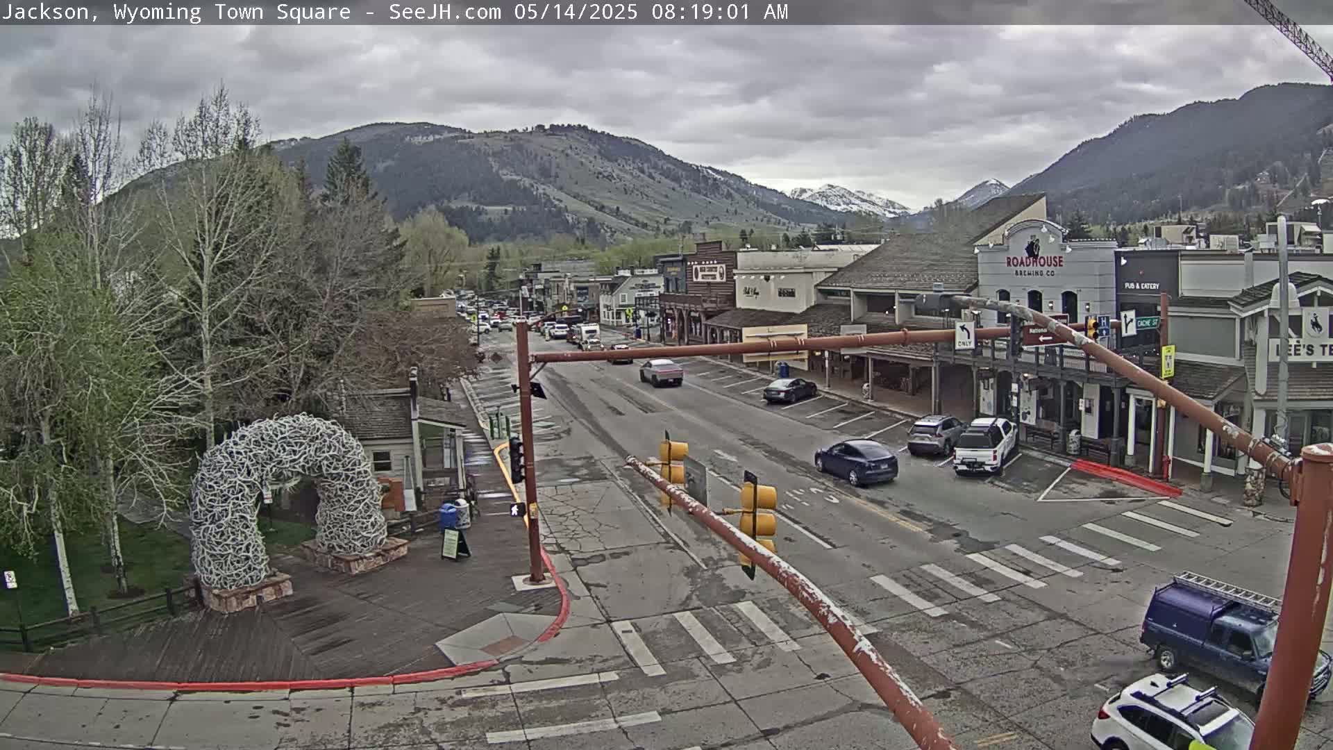 A town square with a street lined with buildings, cars, and a large antler arch is visible under an overcast sky, with mountains in the background.