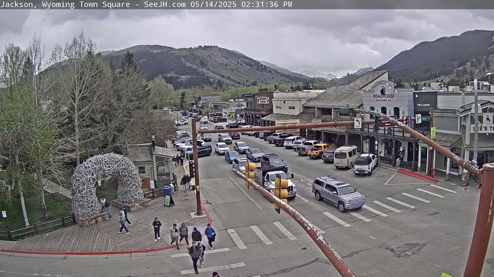 A town square with a large antler arch, numerous parked and moving vehicles, and pedestrians on a partly cloudy day, with mountains in the background.