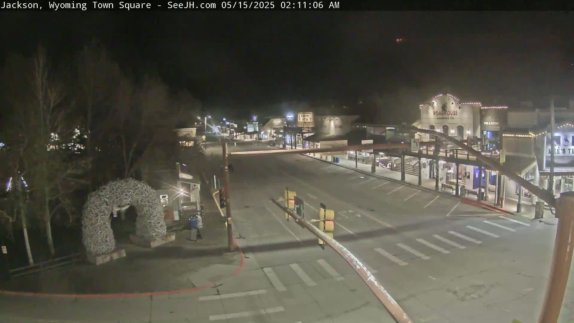An empty town square at night, illuminated by streetlights, features a large antler arch and a few buildings with lights on.