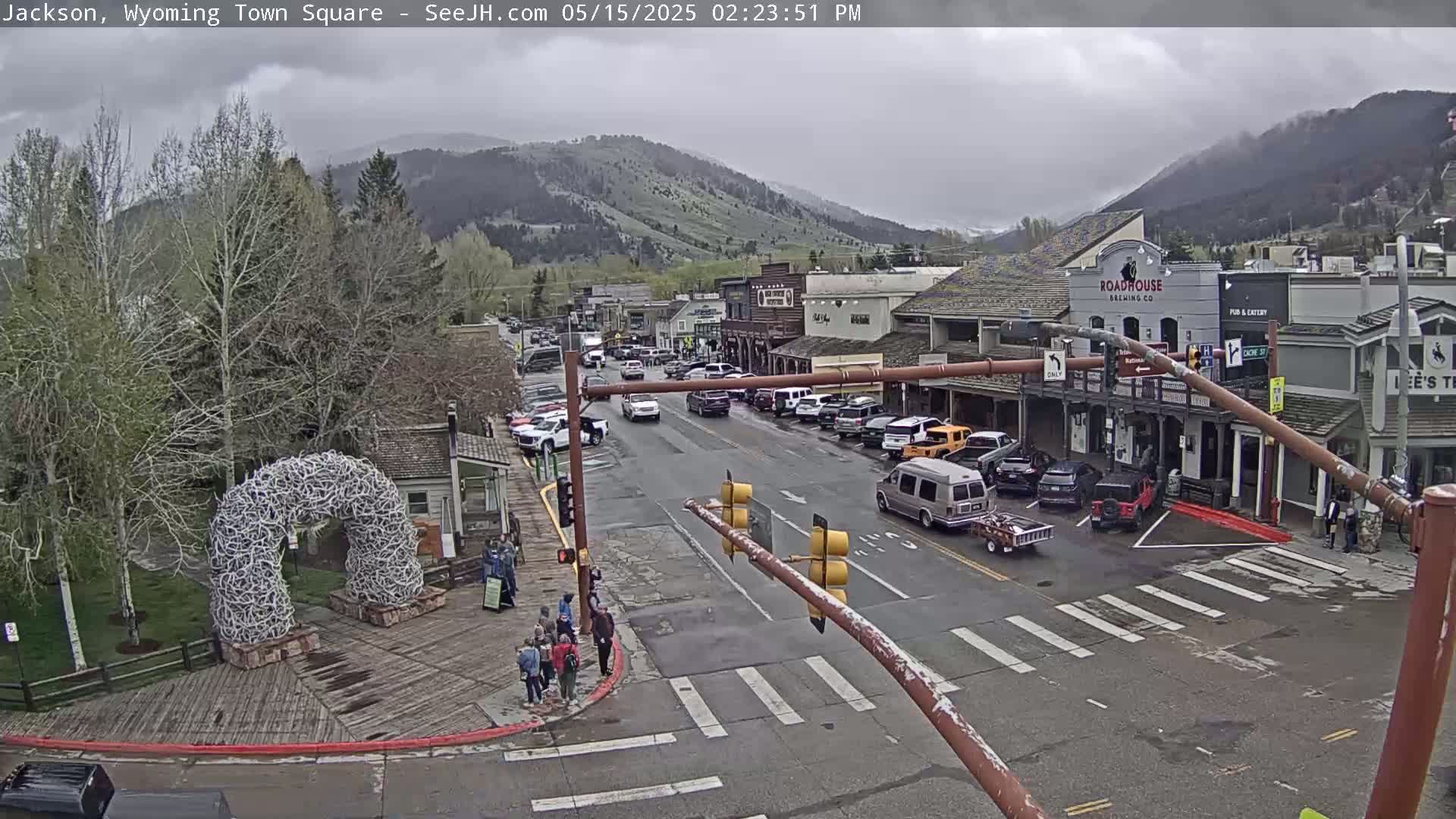 A town square scene on an overcast day shows a street with parked cars and some pedestrians, a large antler arch, and buildings with businesses against a backdrop of mountains.