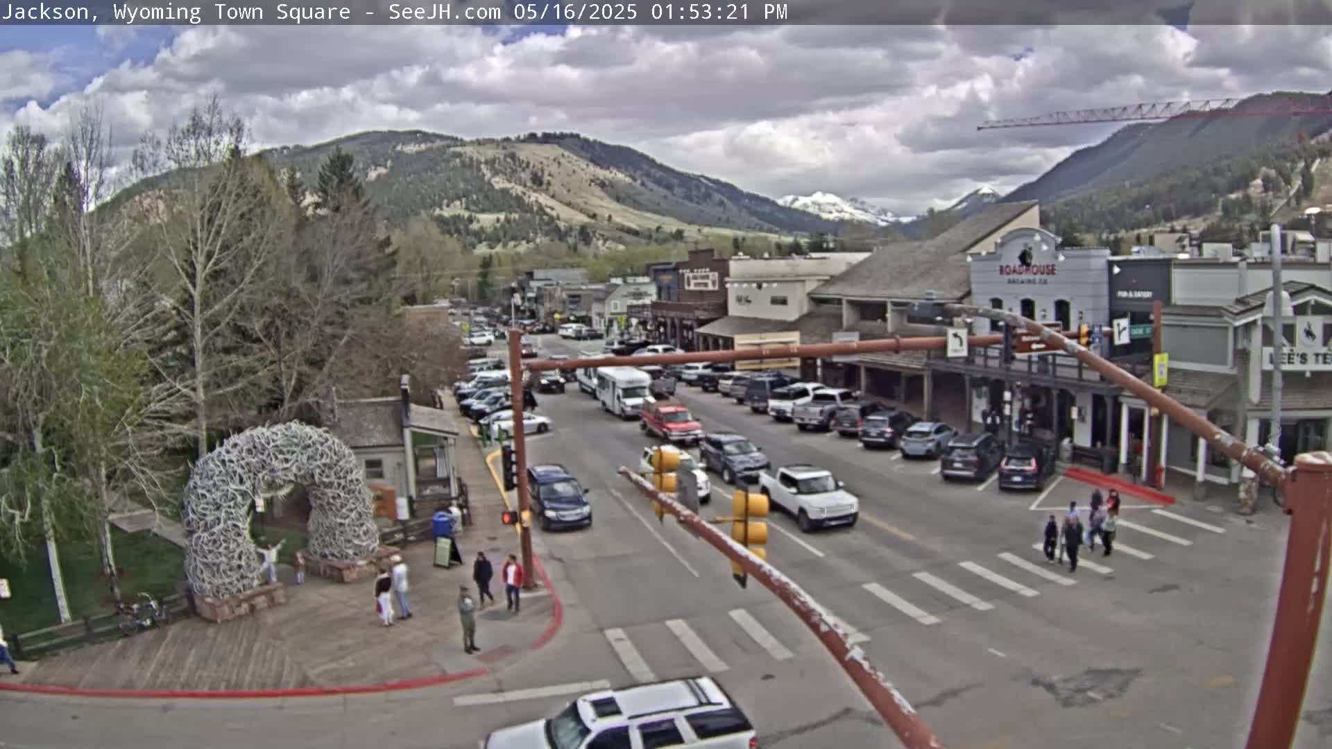 A partly cloudy day reveals a town square with parked cars lining the streets, pedestrians crossing a crosswalk, and a large antler archway near a grassy area, all set against a backdrop of mountains.