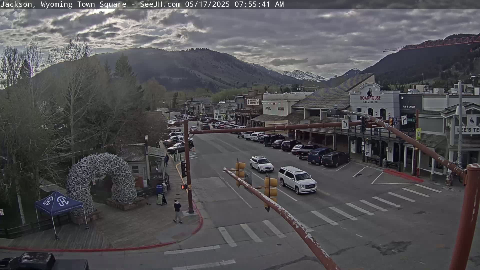 A town square with overcast skies, numerous parked cars lining the streets, and several buildings visible against a mountain backdrop.