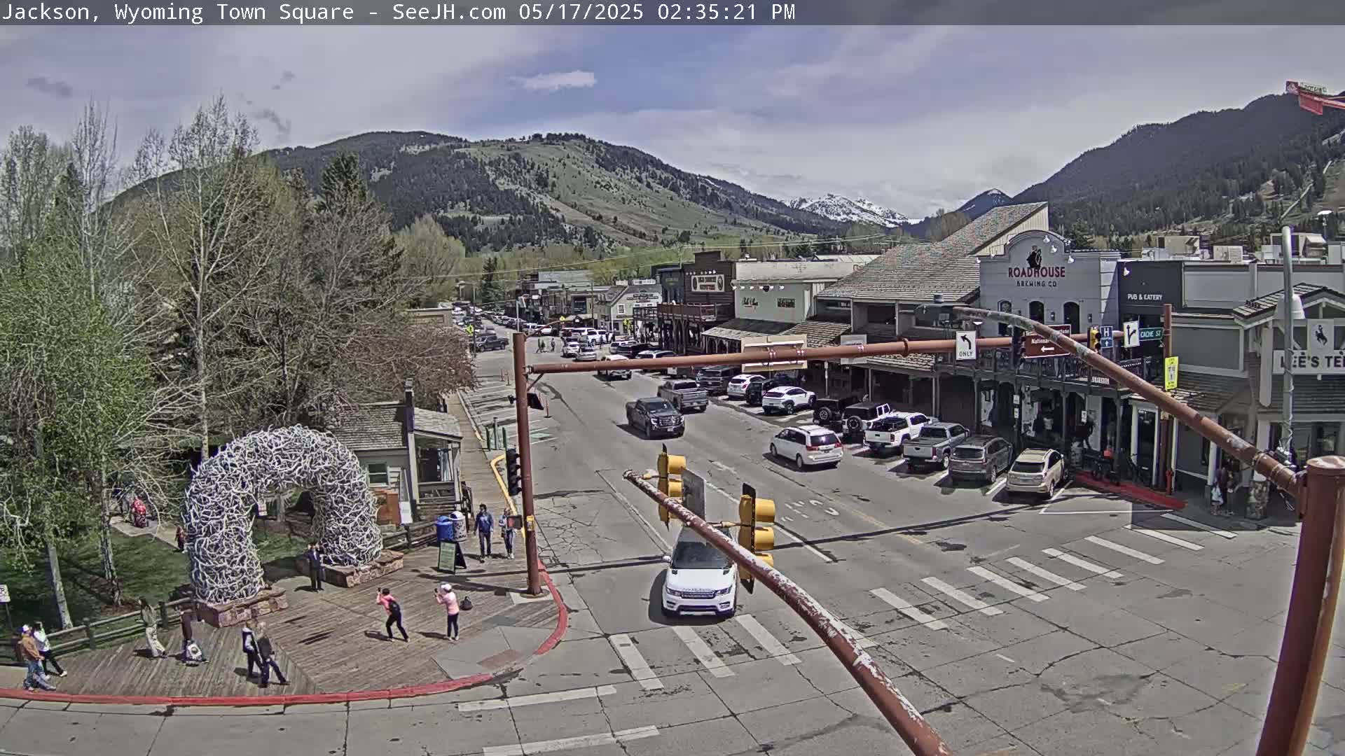 A town square with parked cars and pedestrians, under a partly cloudy sky, is nestled in a valley surrounded by mountains.