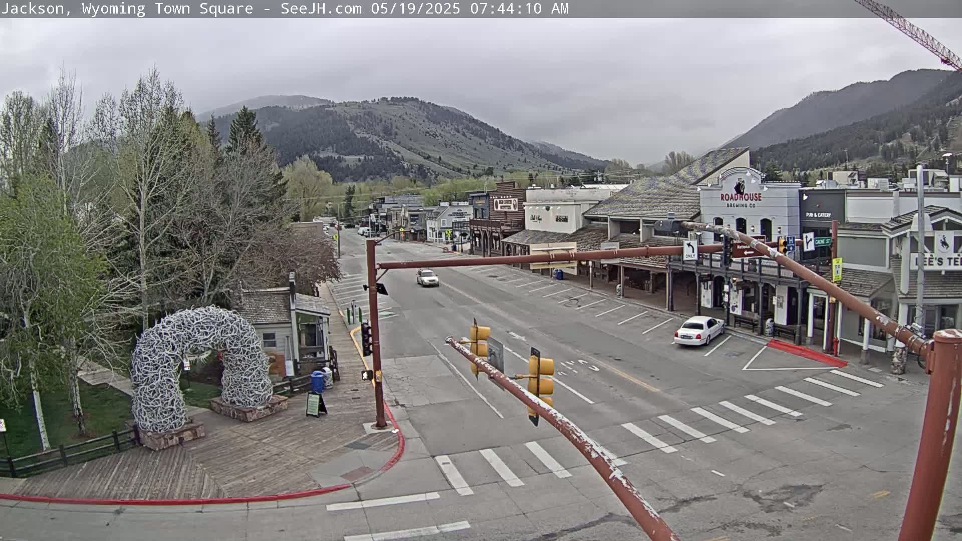 A mostly overcast day reveals a town square with a few cars on the street, buildings lining the sides, and a large antler arch in a nearby park, all set against a backdrop of mountains.