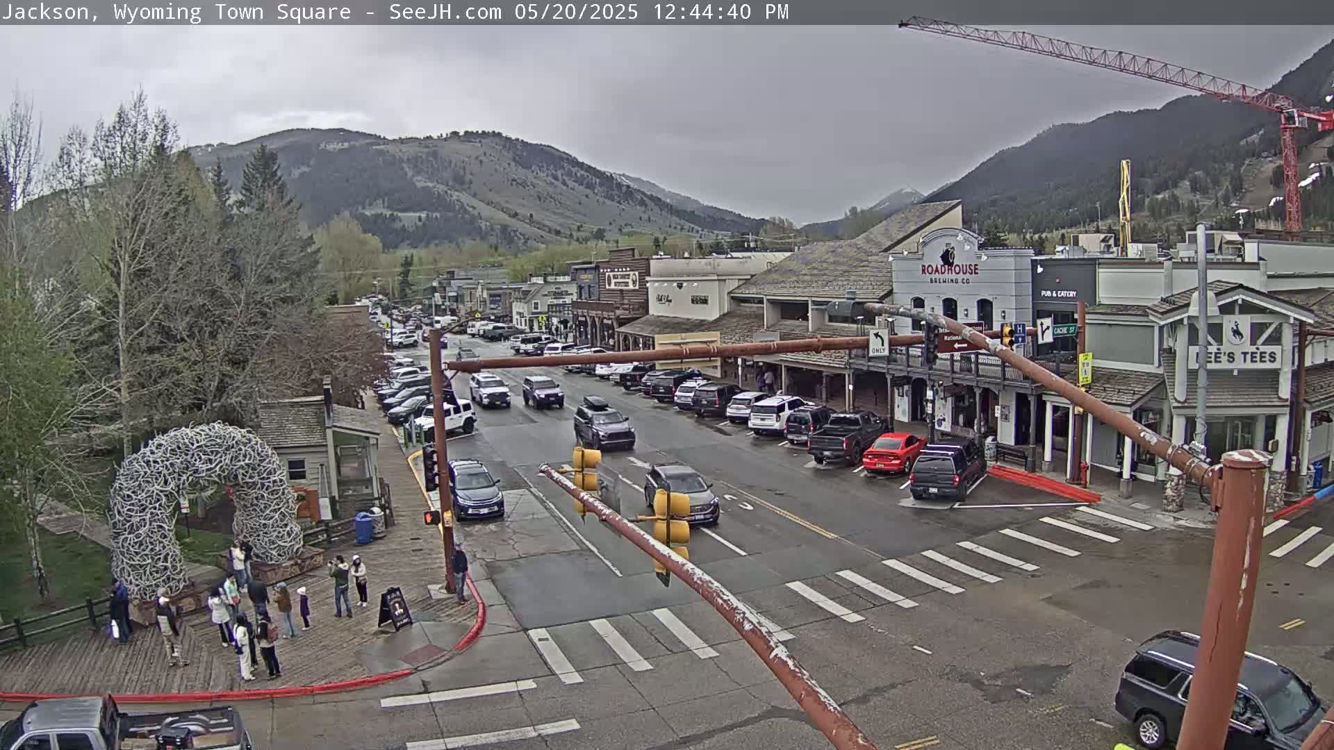 A town square with overcast skies shows a street scene with parked and moving cars, pedestrians, and buildings, including a brewery, against a backdrop of mountains.