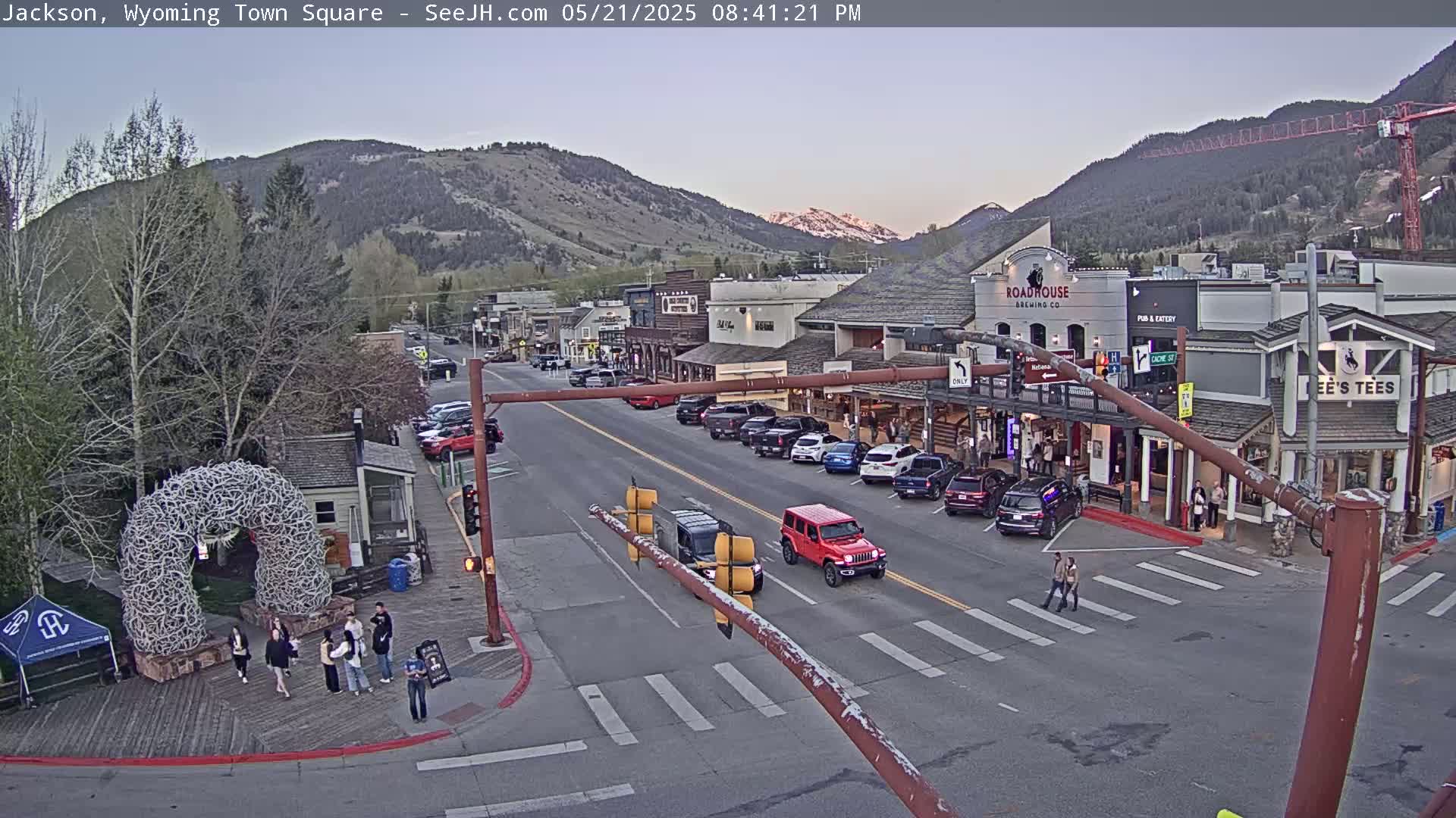 A town square scene at dusk, under a mostly clear sky, shows a street with parked and moving cars, buildings with storefronts, and people walking near a large antler arch.