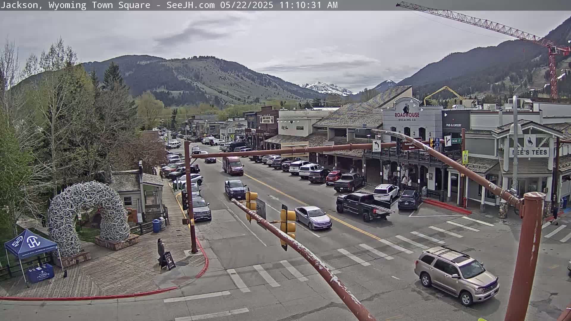 A town square with a street lined with buildings, vehicles, and pedestrians under a partly cloudy sky, with mountains in the background.