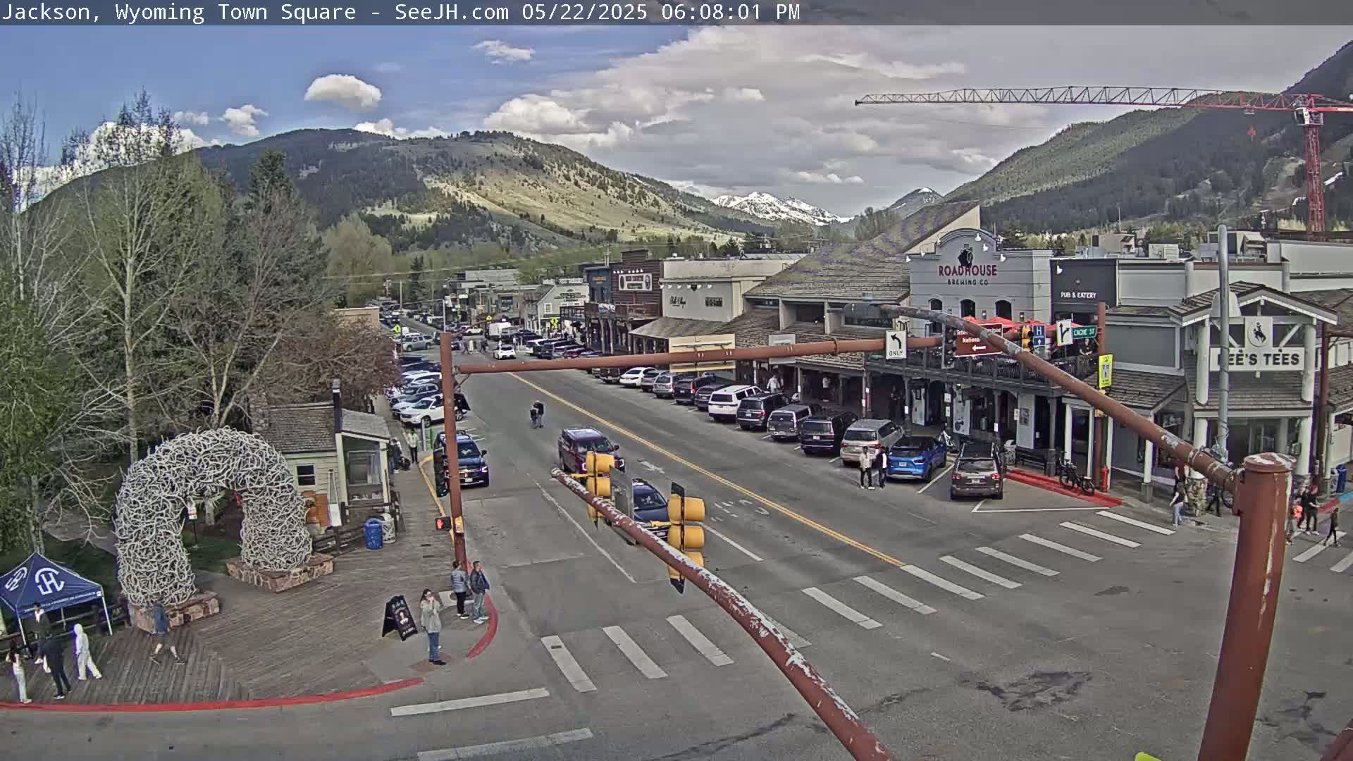 A partly cloudy day in a town square, showing a street with parked cars and pedestrians, buildings lining the street, and mountains in the background.