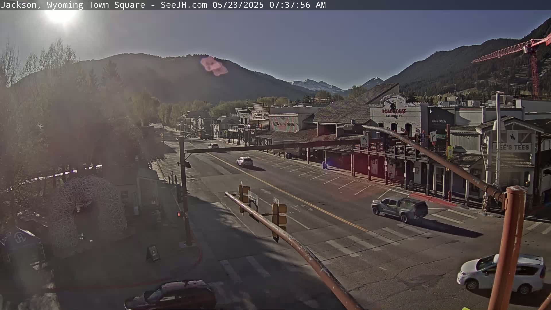 A sunny morning view of a town square with several vehicles on a roadway lined with buildings, and mountains in the background.