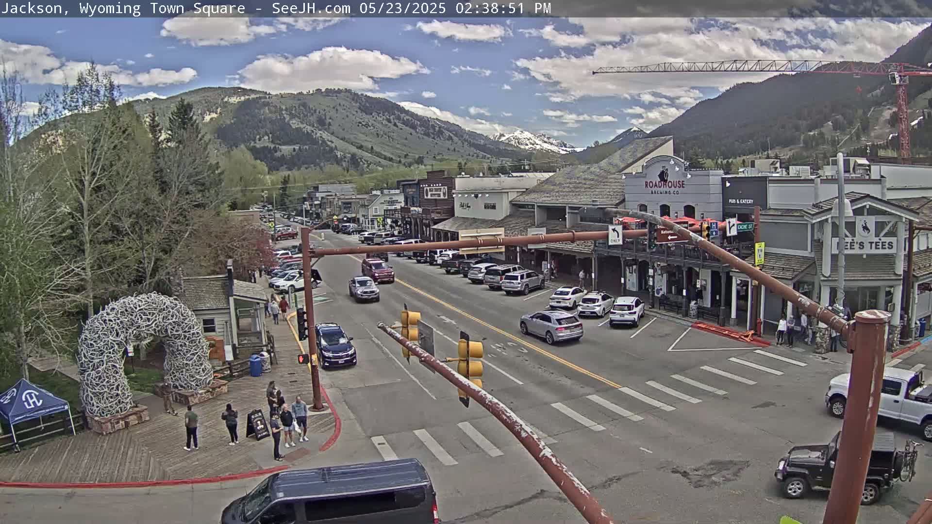 A town square with parked cars and pedestrians, under a partly cloudy sky, is nestled against a backdrop of mountains.