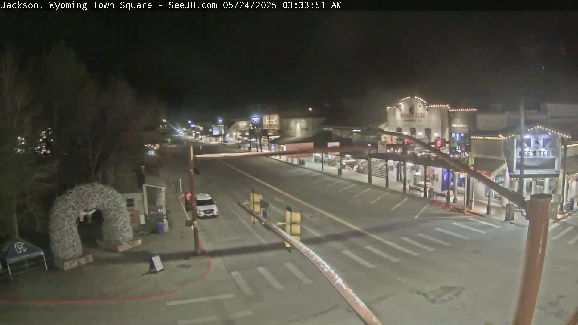A nighttime view of a mostly empty town square with several buildings, a vehicle, and a large antler archway.
