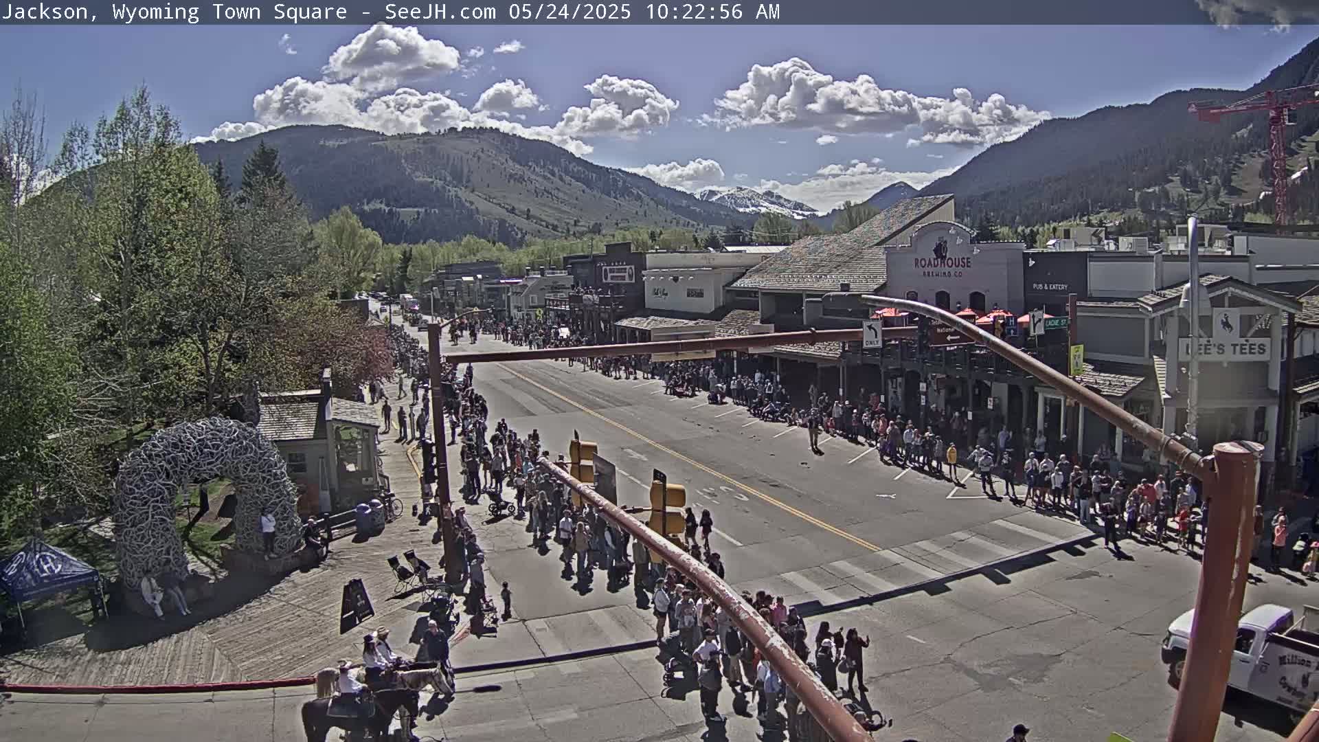 A sunny day shows a large crowd of people lining a town square street, with mountains in the background.