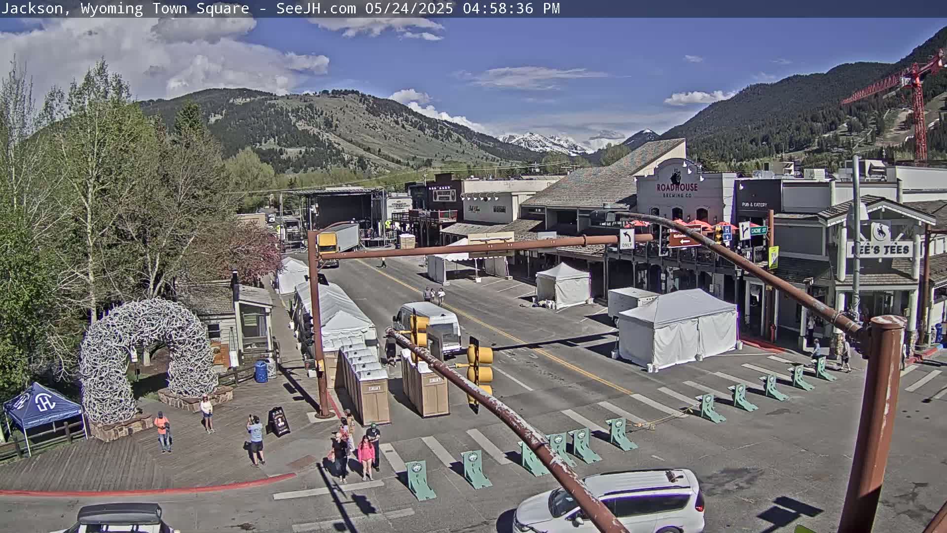 A sunny day in a town square shows several tents and people walking near buildings and a large antler arch, with mountains in the background.